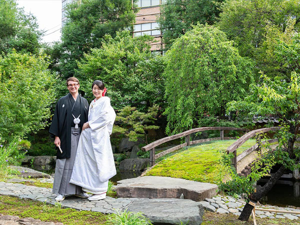 緑豊かな日本庭園で、紋付羽織袴の新郎と白無垢を纏った新婦が笑顔で並ぶ結婚写真