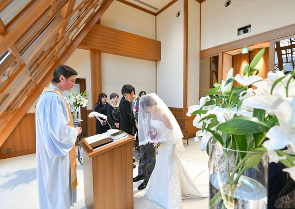 明るいチャペルで神父の前で頭を下げる、ウェディングドレス姿の新婦とタキシード姿の新郎