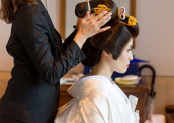 白無垢の新婦が、ヘアメイクスタッフによって伝統的な日本髪（文金高島田）と簪を整えられている支度風景