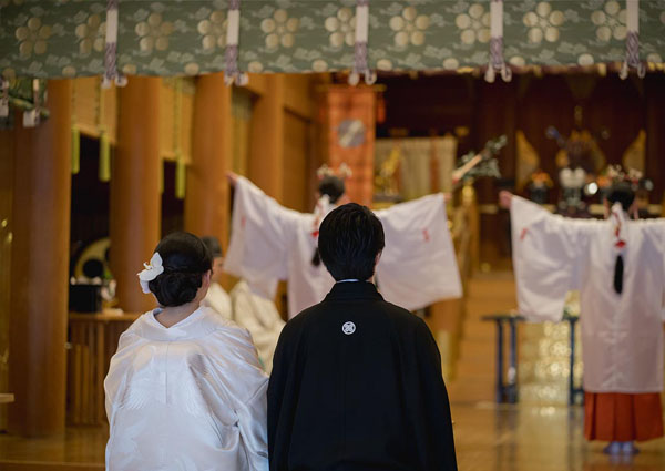 神前式の挙式中、社殿で巫女による神楽舞を静かに見守る白無垢の新婦と紋付袴の新郎の後ろ姿