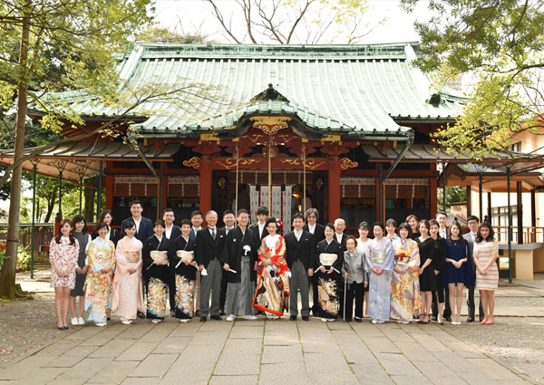 神社の社殿を背景にした、色打掛姿の花嫁と新郎、参列者の集合写真。