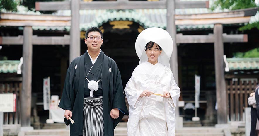 由緒ある神社の社殿と鳥居を背景に、綿帽子を被った白無垢姿の花嫁と、黒の紋付袴を凛々しく着こなした新郎が並んで立つ、神前式の記念ポートレート。