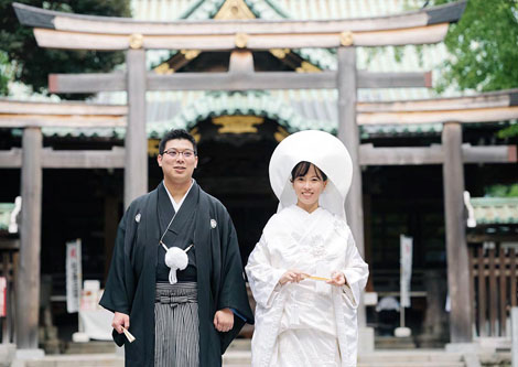 神社の鳥居と社殿を背景に、綿帽子姿の白無垢の花嫁と紋付袴の新郎の記念写真。