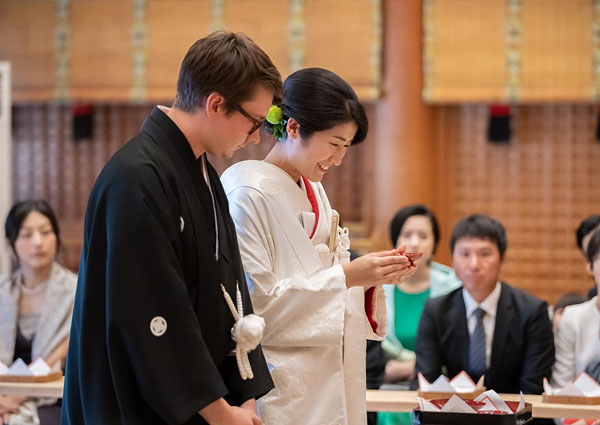 神前式で向き合う眼鏡の新郎と白無垢の新婦、見守る参列者